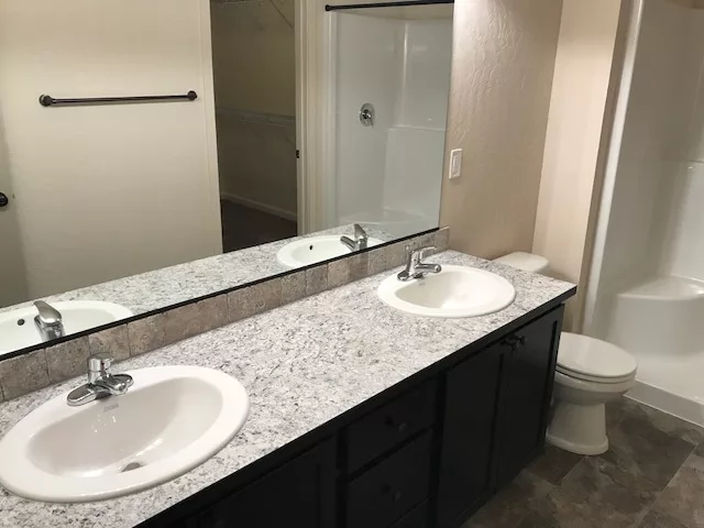 Double-sink bathroom vanity with granite countertop, large mirror, and adjacent shower and toilet.