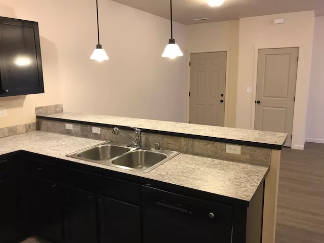 Kitchen with dark cabinets, a dual sink, granite countertops, and pendant lights, facing an open doorway.