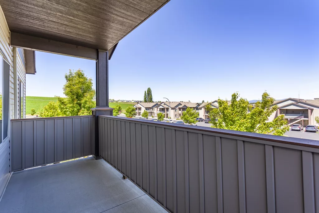 A balcony with a wooden railing overlooks a residential neighborhood with clear skies and leafy trees.