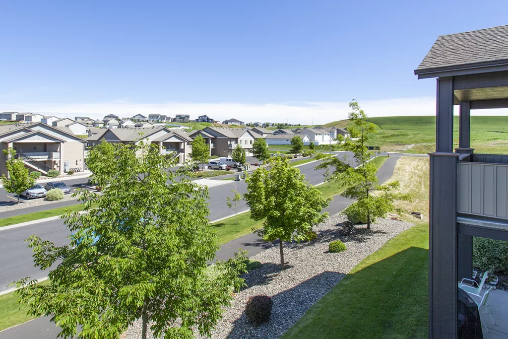 Suburban neighborhood with tree-lined streets, houses, and a distant grassy hill under a clear blue sky.