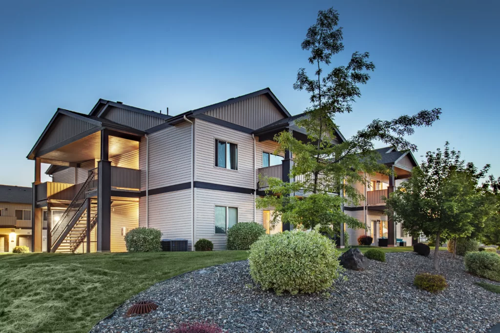 Apartment building with a landscaped yard featuring bushes and small trees under a clear evening sky.