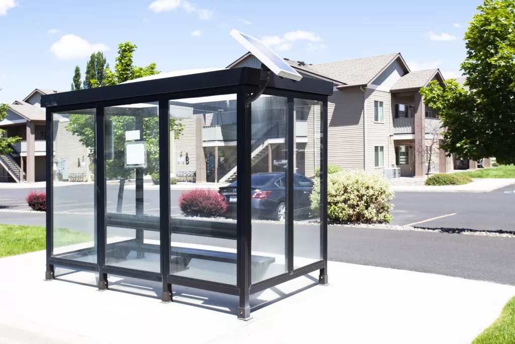 A glass bus shelter with a solar panel on top, situated in front of a residential building on a sunny day.