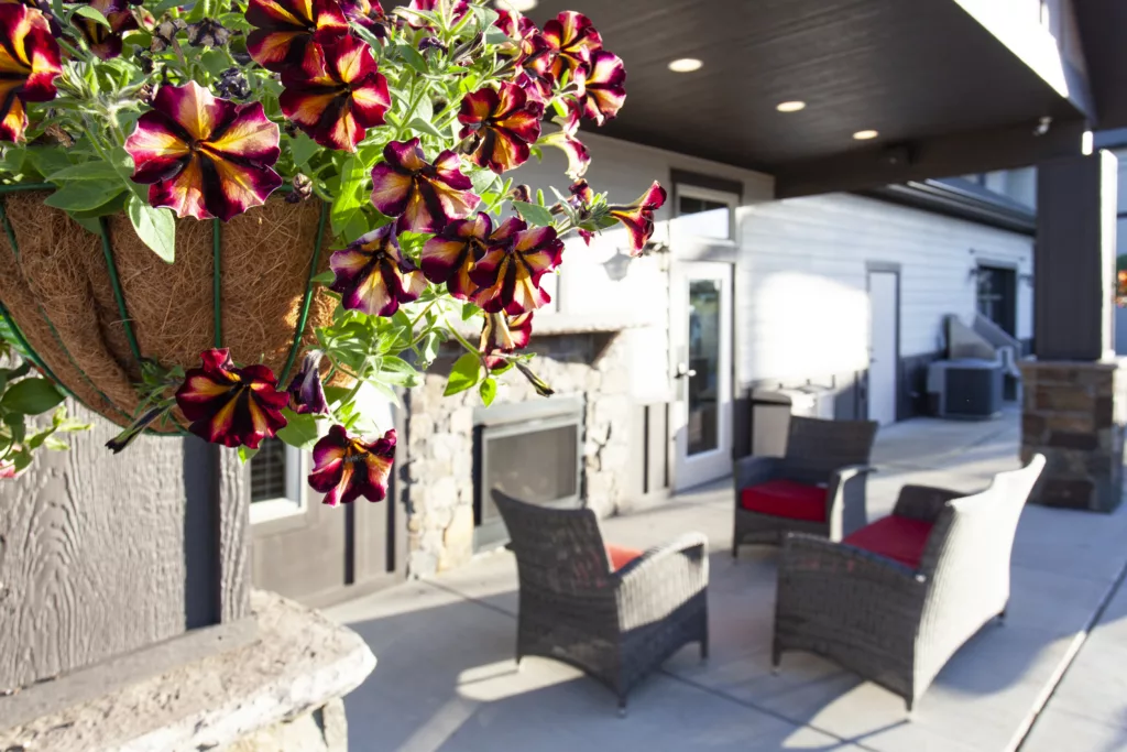 Hanging basket with vibrant flowers on a patio, with wicker chairs and a fireplace in the background.
