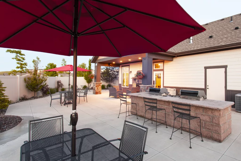 Patio with a red umbrella over a table, metal chairs, and a stone outdoor kitchen with grills beside a house.