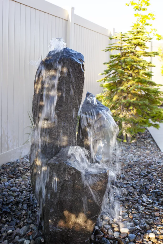Water flowing over three stacked rocks in a landscaped garden with a small evergreen tree nearby.
