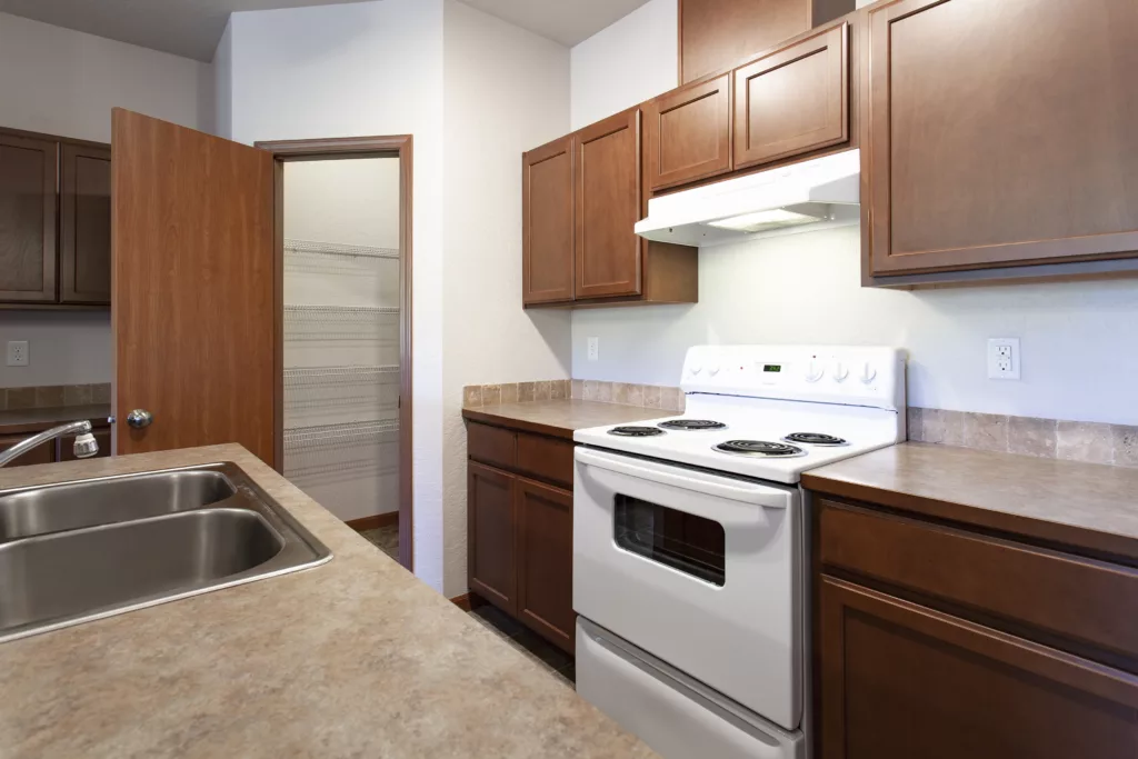 A kitchen with brown wood cabinets, an electric stove, a range hood, and a double sink on a marble countertop.