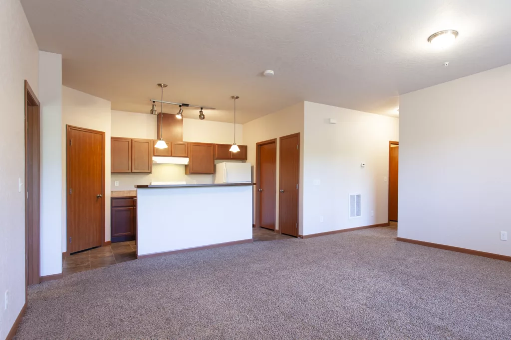 An empty apartment with beige walls, carpeted living area, and a kitchen with wooden cabinets and pendant lighting.