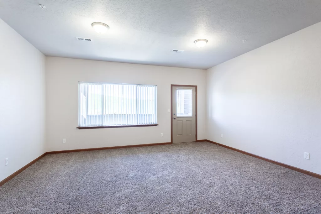 An empty room with carpeted floors, white walls, a window with vertical blinds, and a door leading outside.