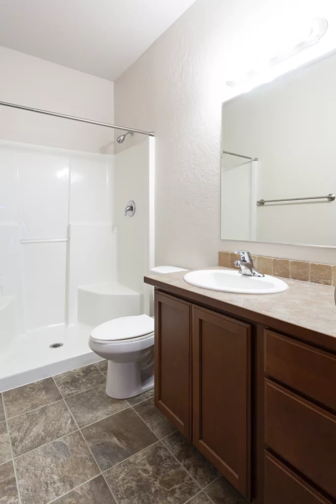 Bathroom with a white shower, toilet, and wooden vanity with a sink and mirror; features brown tiled floor.
