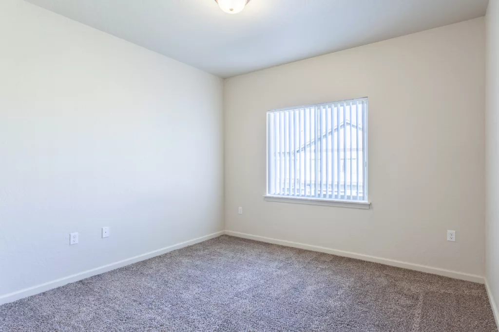 Empty room with beige walls, carpeted floor, and a window with vertical blinds.