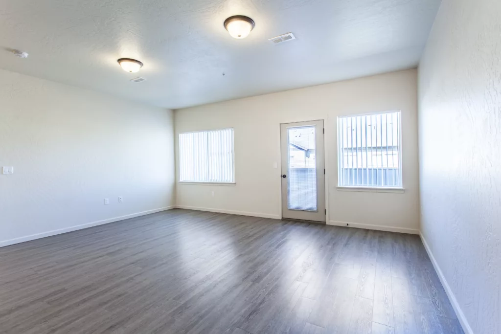 An empty room with wood flooring, two windows with blinds, and a glass door leading outside.