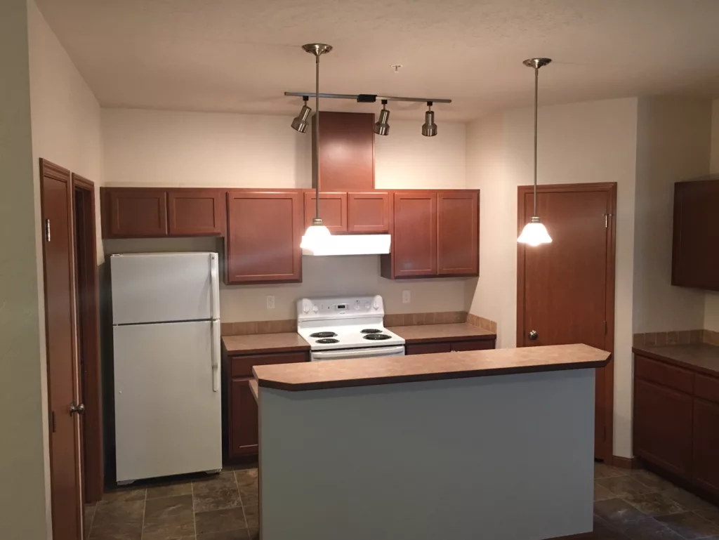A kitchen with brown cabinets, a white refrigerator, an oven, and overhead lighting above a small island.